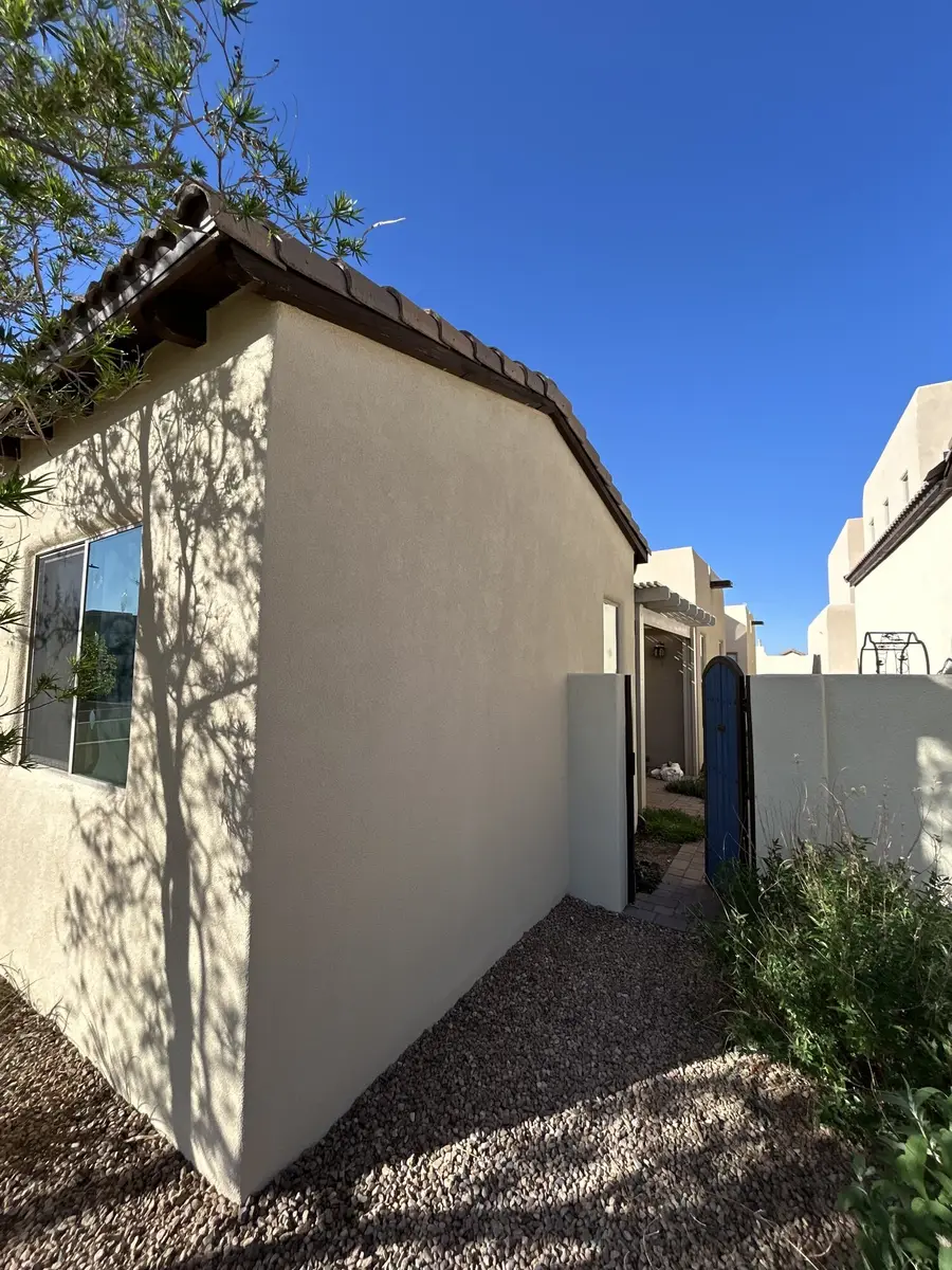 Cream stucco casita wall under a clay tile roof, with tree shadows across it and a blue gate to one side.