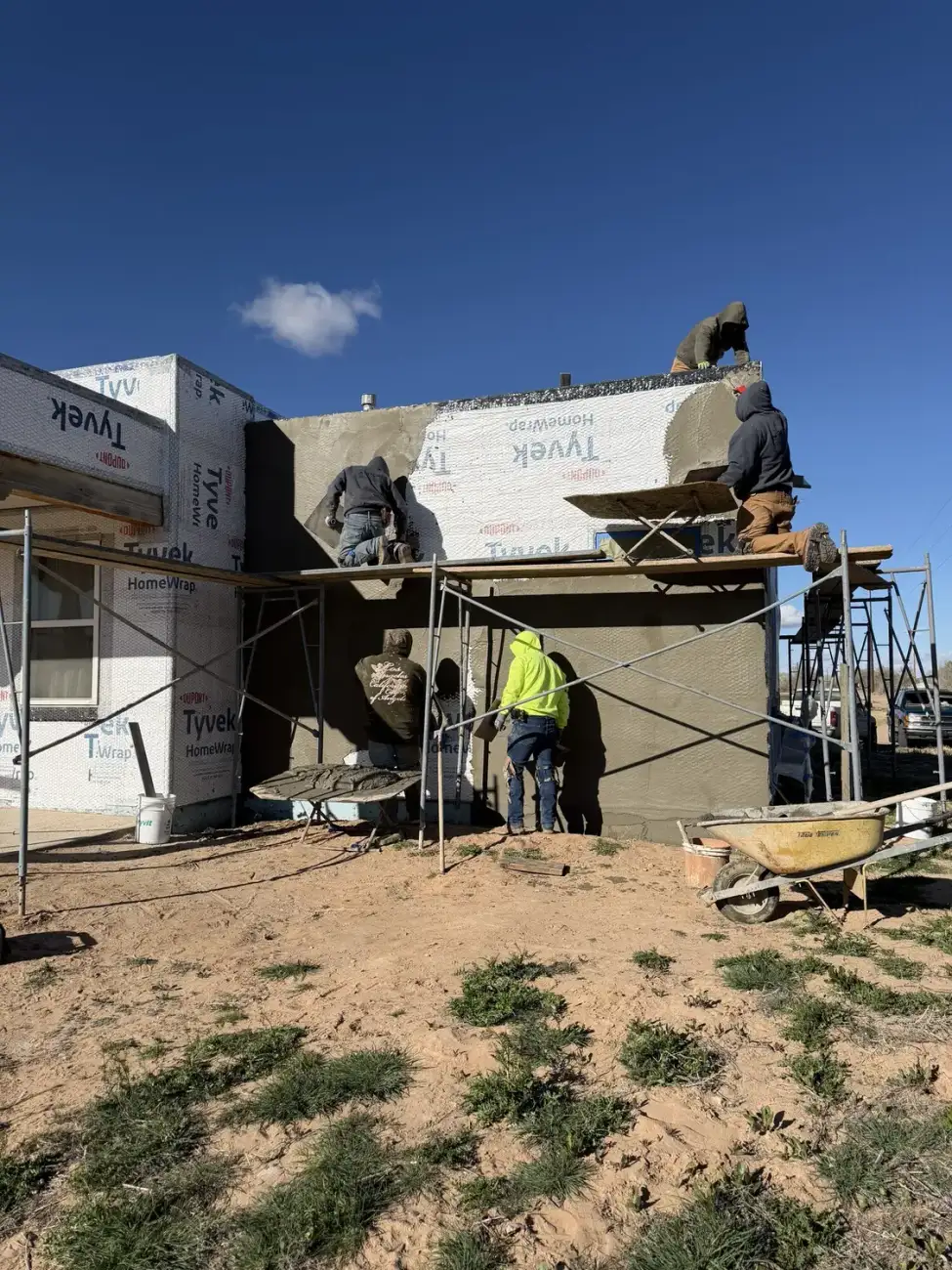 Crew on scaffolding troweling a grey stucco base coat onto a house wall over housewrap and lath.