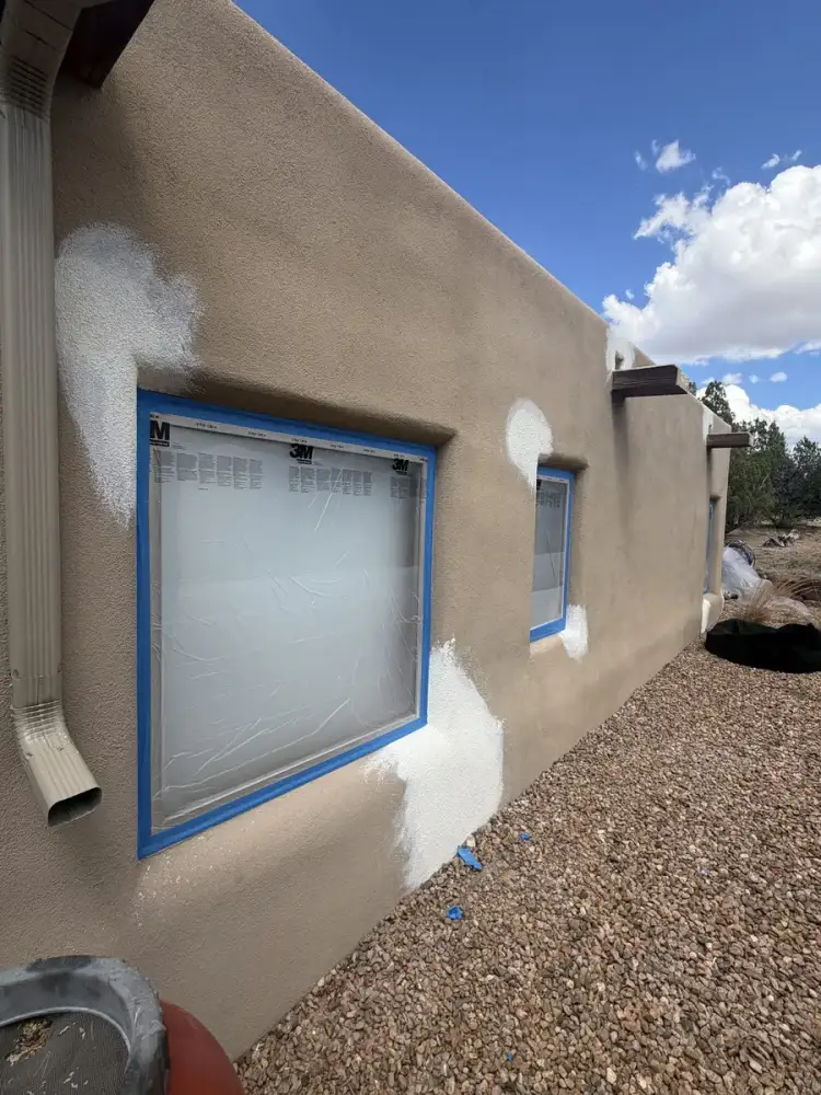Tan stucco wall with white patches around the windows, the glass masked off in plastic and blue tape.