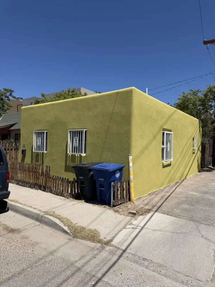 Corner of a single story Albuquerque house finished in a yellow green stucco color coat, with barred windows and a wooden fence.
